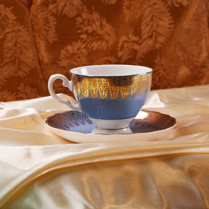 Op de afbeelding: Een blauw en goud theekopje en schotel set. Het theekopje heeft een blauwe body met gouden filigraan rond de rand. De schotel is wit met een gouden rand. De set staat op een cr&egrave;mekleurig satijnen oppervlak.