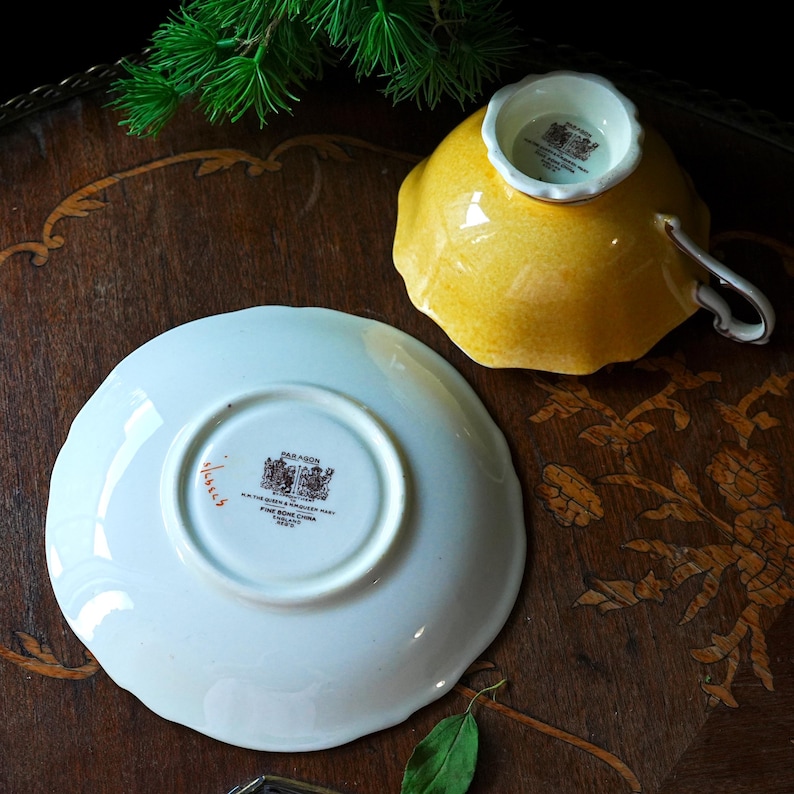 May include: A vintage yellow teacup and saucer set. The teacup is upside down, showing a white interior. The saucer is white with a scalloped edge and a Paragon stamp. The set is displayed on a dark wooden surface with decorative inlay.