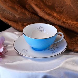 Pode incluir: Um conjunto de ch&aacute;vena e pires azul claro. A ch&aacute;vena tem uma borda dourada e um interior branco. O pires &eacute; branco com uma borda dourada e um desenho floral. O conjunto est&aacute; sobre uma superf&iacute;cie de cetim branco.