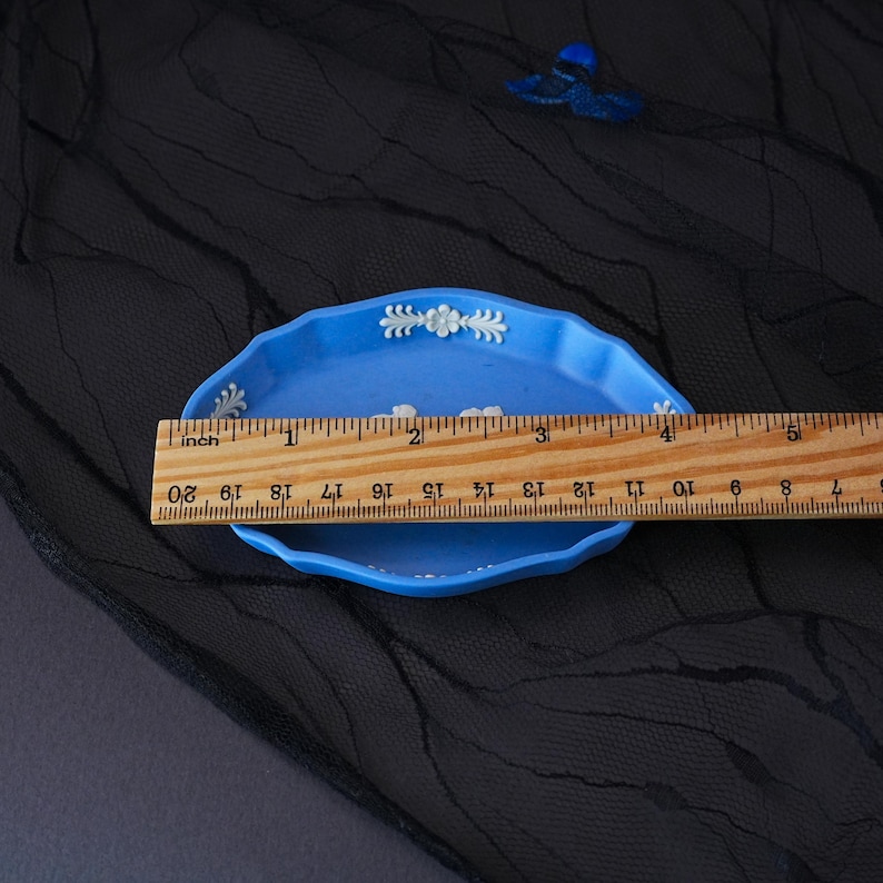 May include: A light blue oval ceramic dish with white floral relief detailing along the scalloped rim. A wooden ruler is placed across the dish, showing a width of approximately 11.5 cm. The dish features a central white relief design.