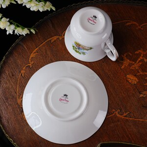 May include: A white teacup and saucer set with a landscape design and gold trim. The teacup is resting on the saucer, which is also white. The set is on a wooden surface. The text "Queen Anne" is visible.