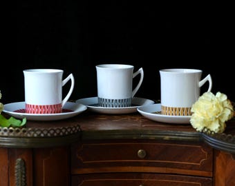 May include: Three white ceramic coffee cups with matching saucers. Each cup has a different geometric pattern in red, blue, and gold around the base. The cups and saucers are on a wooden surface with a dark background.