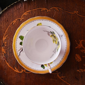 Peut inclure: Service &agrave; th&eacute; blanc avec motif floral jaune et vert et bordure dor&eacute;e. La tasse repose sur une soucoupe assortie, le tout pos&eacute; sur une table en bois fonc&eacute;. L'ensemble est con&ccedil;u pour le th&eacute; ou le caf&eacute;.