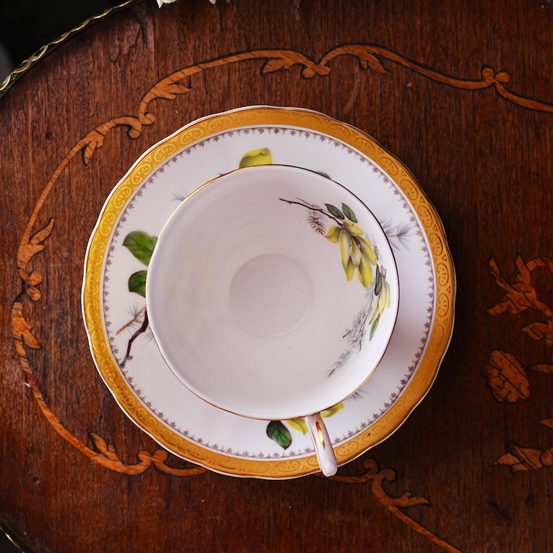 Peut inclure: Service &agrave; th&eacute; blanc avec motif floral jaune et vert et bordure dor&eacute;e. La tasse repose sur une soucoupe assortie, le tout pos&eacute; sur une table en bois fonc&eacute;. L'ensemble est con&ccedil;u pour le th&eacute; ou le caf&eacute;.