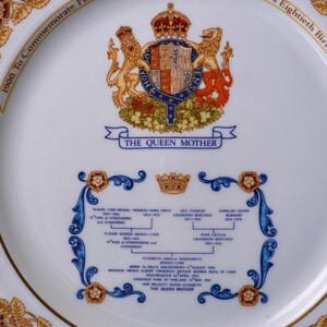 Pode incluir: Um prato comemorativo de porcelana branca com um bras&atilde;o real colorido com le&otilde;es e uma coroa, o texto THE QUEEN MOTHER e uma &aacute;rvore geneal&oacute;gica azul detalhada com detalhes florais dourados ao longo da borda do prato.