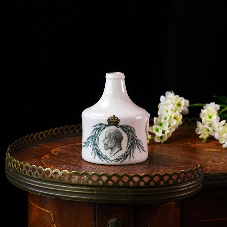 K&ouml;nnte beinhalten: Wei&szlig;e Keramikflasche mit einem Portr&auml;t eines Mannes, einer Krone und Lorbeerbl&auml;ttern. Die Flasche hat einen breiten Boden und einen schmalen Hals. Das Portr&auml;t ist in Schwarzwei&szlig;, die Bl&auml;tter sind gr&uuml;n.