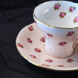 Może przedstawiać: Biały zestaw filiżanki do herbaty ze spodkiem wykonany z porcelany, ozdobiony delikatnym wzorem małych r&oacute;żowych r&oacute;ż z zielonymi liśćmi. Zar&oacute;wno filiżanka, jak i spodek wykończone są cienkim złotym obrzeżem na falowanych krawędziach, co nadaje im elegancji.