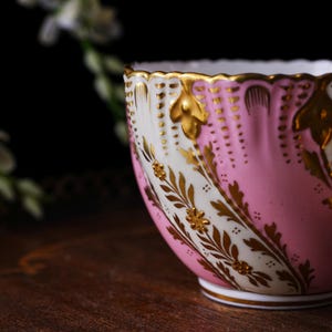 May include: A close-up of a pink and white teacup with gold accents. The teacup features a scalloped rim and is decorated with gold floral patterns. The cup sits on a white base and has a gold trim.