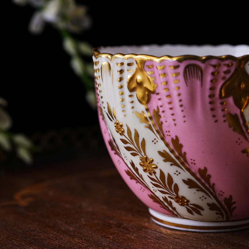 May include: A close-up of a pink and white teacup with gold accents. The teacup features a scalloped rim and is decorated with gold floral patterns. The cup sits on a white base and has a gold trim.