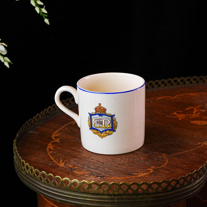 Op de afbeelding: Witte keramische mok met een blauwe rand en oor. De mok heeft een wapen met een kroon en de letters "ER" in blauw en goud. De mok staat op een houten tafel.