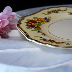Puede incluir: Un plato crema con borde festoneado y un dise&ntilde;o floral con flores amarillas, rojas y moradas. El plato tiene un borde dorado y un borde decorativo. Una flor rosa est&aacute; en el fondo.