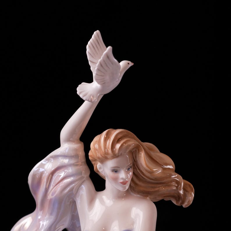 May include: A porcelain figurine of a woman with flowing auburn hair, holding a white dove aloft. The woman is wearing a flowing gown with iridescent accents. The sculpture is set against a black background.