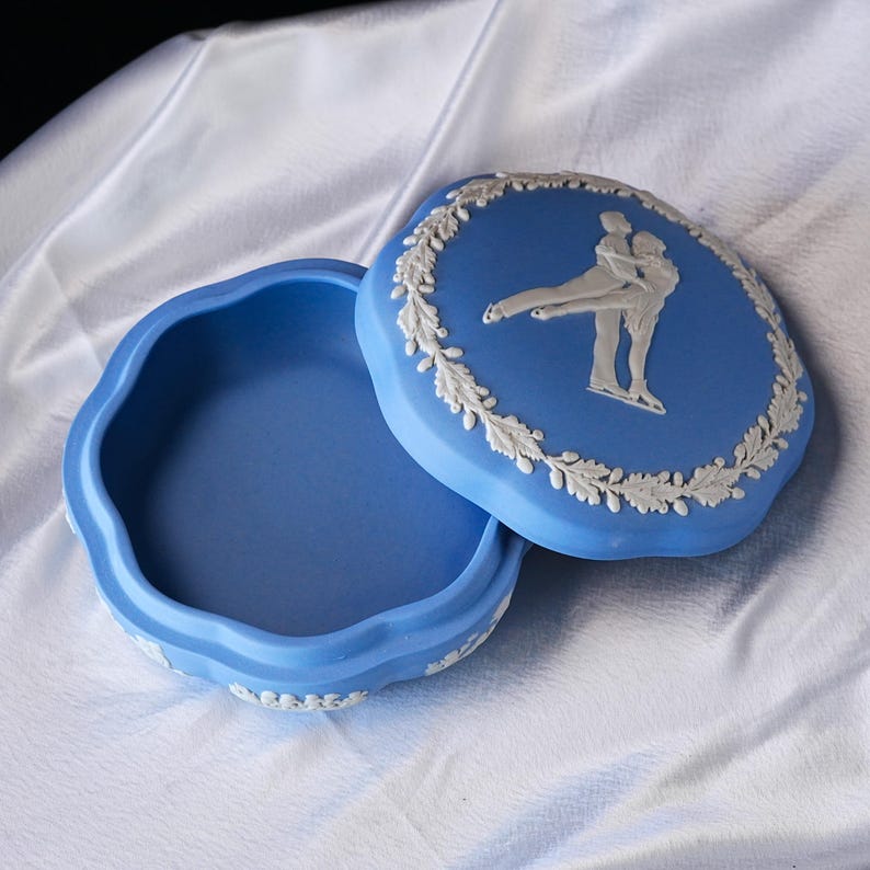 Może przedstawiać: Niebieskie ceramiczne pudełko na drobiazgi z pokrywką przedstawiającą biały relief łyżwiarzy figurowych. Pudełko ma falisty brzeg i białą girlandę z liści wok&oacute;ł krawędzi pokrywki. Wnętrze pudełka jest r&oacute;wnież niebieskie.