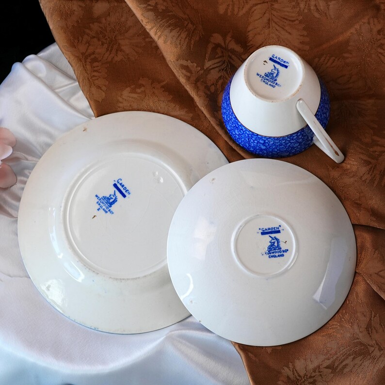 May include: A white teacup with a blue speckled pattern and a matching saucer and plate. The teacup and saucer have a blue and white floral design. The plate and saucer have the text "GARDEN" and "WEDGWOOD ENGLAND" in blue.