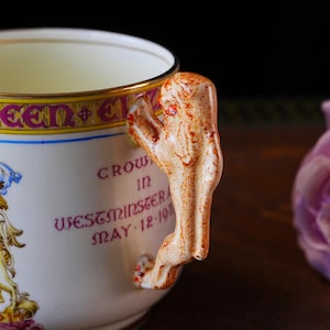 Puede incluir: Taza de t&eacute; color crema con un asa decorativa. La taza presenta un borde dorado y el texto "CROWN IN WESTMINSTER MAY 12-1937". El asa tiene forma de figura con detalles en naranja y marr&oacute;n.