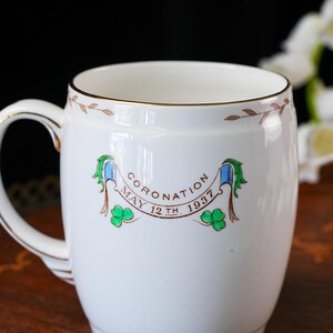 Może przedstawiać: Biały ceramiczny kubek ze złotym obrzeżem i brązowym wzorem liści wok&oacute;ł g&oacute;rnej krawędzi. Na środku znajduje się baner z napisem CORONATION MAY 12TH 1937, otoczony niebiesko-zielonymi wstążkami oraz ilustracjami zielonych koniczynek na białym tle.