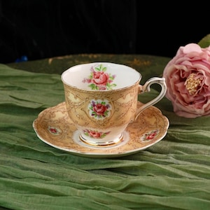 May include: A vintage teacup and saucer set. The cup and saucer are a light tan color with a floral design in pink and red. The cup has a curved handle and a gold rim. The saucer has a scalloped edge.