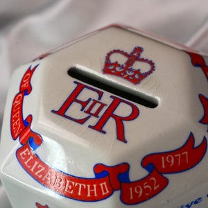 Op de afbeelding: Een witte keramische spaarpot met een gleuf voor munten. De spaarpot heeft de tekst "ER" in het rood, een rode kroon en rode banners met de tekst "ELIZABETH" en de jaren "1952" en "1977".