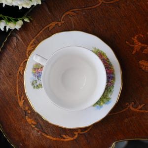 May include: A white teacup and saucer set with a floral design and gold trim. The teacup sits on a matching saucer, both placed on a wooden surface. The floral design includes shades of pink, green, and blue.
