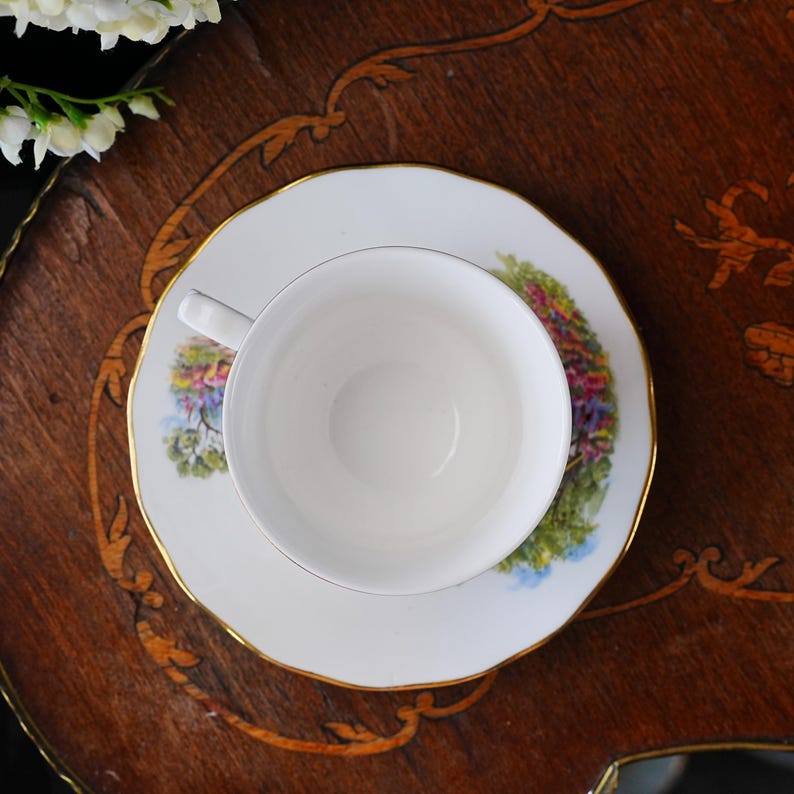 May include: A white teacup and saucer set with a floral design and gold trim. The teacup sits on a matching saucer, both placed on a wooden surface. The floral design includes shades of pink, green, and blue.