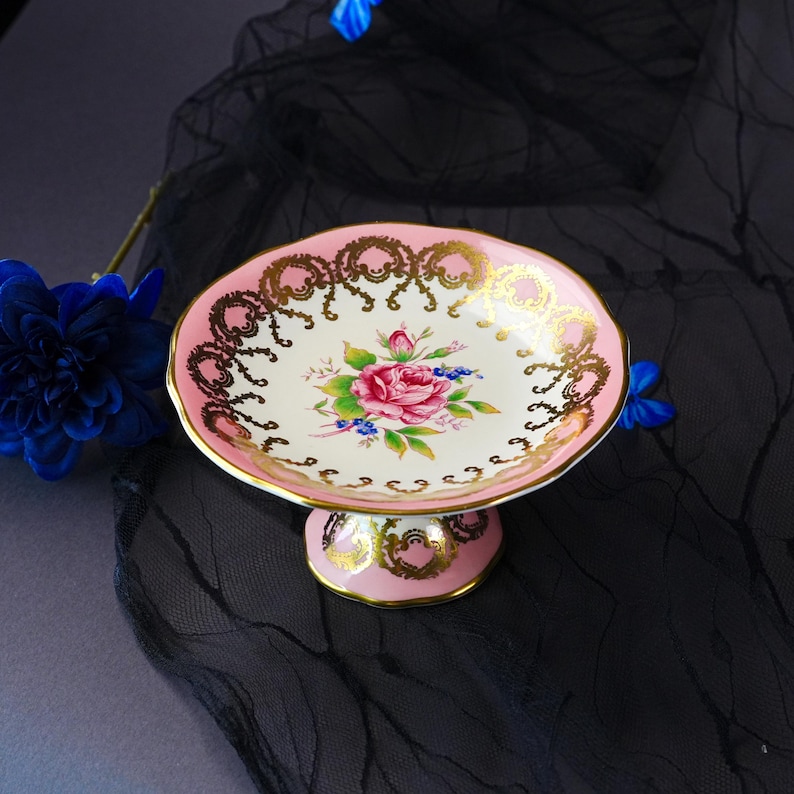 Puede incluir: Un cuenco con pedestal que presenta un centro de color crema con una ilustraci&oacute;n de una rosa rosa y hojas verdes. El borde y la base son de color rosa con detalles de filigrana dorada. El cuenco tiene un borde festoneado y una base circular.