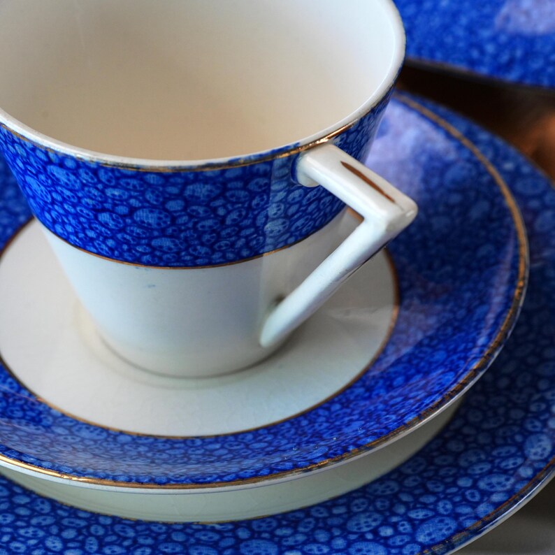 May include: A white teacup, saucer, and plate set with a blue speckled pattern and gold trim. The teacup has a white handle with gold accents. The set is designed for serving tea or coffee.