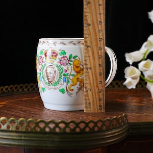 Może przedstawiać: Biały ceramiczny kubek z portretem kr&oacute;la Edwarda VIII, koroną i lwem. Drewniana linijka przystawiona do kubka wskazuje wysokość około 10 cm. Kubek posiada wygięte ucho oraz złote wykończenie krawędzi i g&oacute;rnej części naczynia.