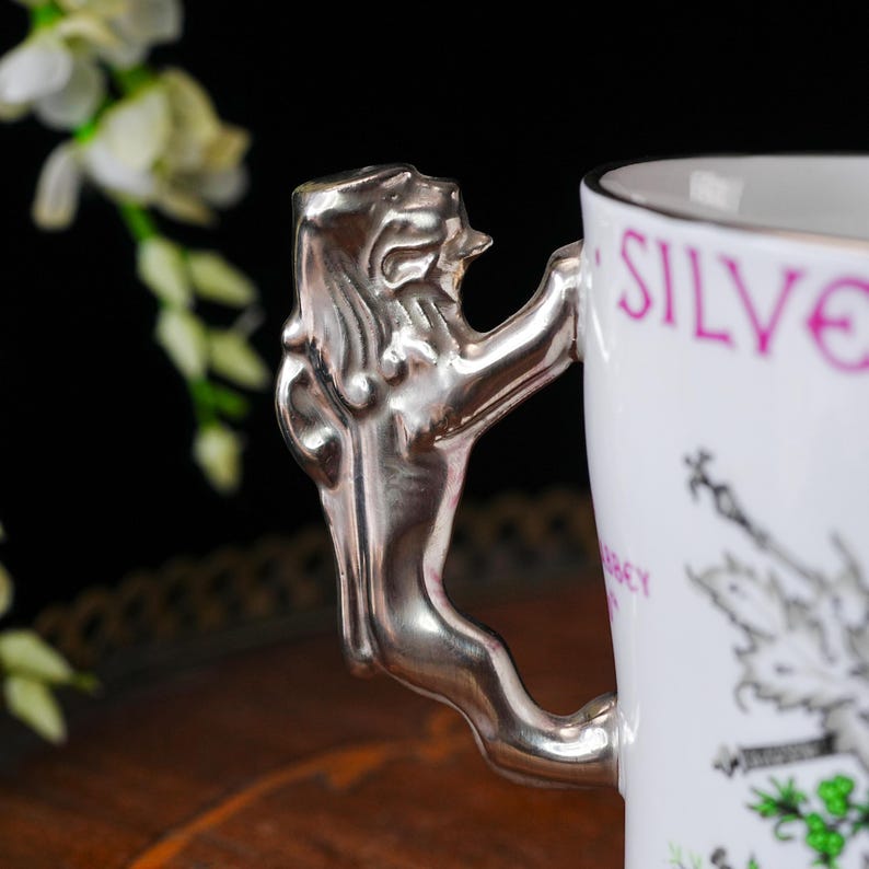 May include: White ceramic mug with a silver lion-shaped handle. The mug has a silver rim and features the word "SILVE" in pink, along with a black and white illustration. The handle is a detailed sculpture of a lion.