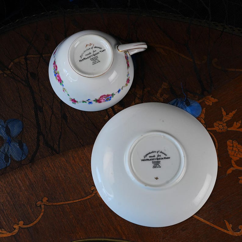 Op de afbeelding: Een witte porseleinen theekop en schotel set met een delicaat bloemmotief van roze en blauwe bloemen. De onderkant van beide stukken toont zwarte tekst: Reproduction of Service made for HER MAJESTY QUEEN MARY, PARAGON FINE BONE CHINA, MADE IN ENGLAND.