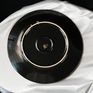 May include: A black ceramic plate with a glossy finish. The plate has a wide rim and a central recessed area. A gold-colored emblem is visible in the center. The plate is likely for decorative or dining use.
