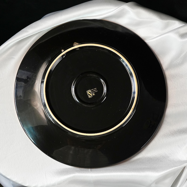 May include: A black ceramic plate with a glossy finish. The plate has a wide rim and a central recessed area. A gold-colored emblem is visible in the center. The plate is likely for decorative or dining use.