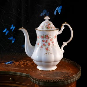 Puede incluir: Una tetera de porcelana blanca con un delicado patrón floral de pequeñas rosas rosadas y hojas verdes. La tetera presenta una base festoneada, un asa curva, un pico largo y una tapa a juego, todo acentuado con un fino borde dorado.