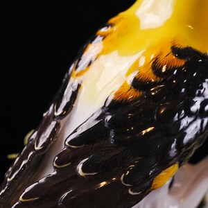 May include: Close-up of a ceramic bird figurine. The bird's feathers are detailed with yellow, black, and white paint. The figurine is on a white branch, with a black background. The bird's wing feathers are a dark brown color.
