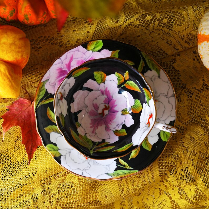 May include: A black teacup and saucer set with a floral design. The teacup and saucer feature a black background with large white and pink peony flowers and green leaves. The set has a gold trim and is on a yellow lace tablecloth.