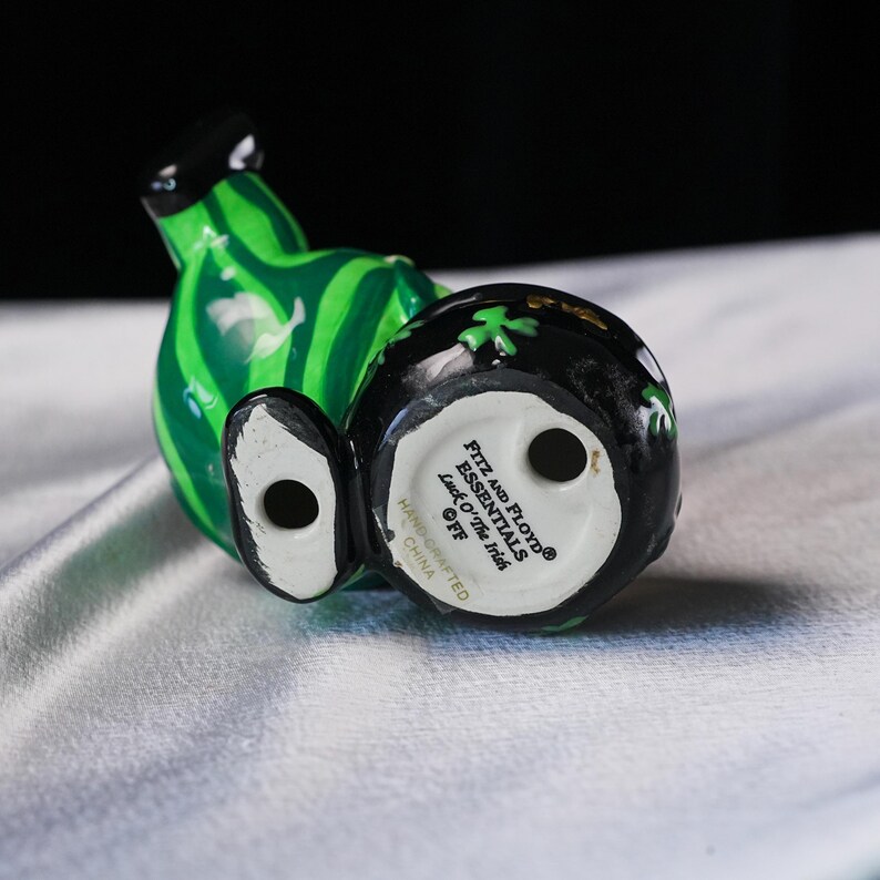 May include: A ceramic figurine of a leprechaun, with a green watermelon-striped body and a black hat. The figurine's head is black with green shamrocks. The base of the figurine has the text "FITZ AND FLOYD ESSENTIALS Luck O' The Irish".