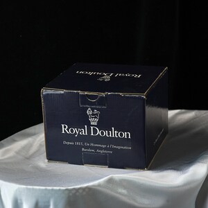 Może przedstawiać: Granatowe kartonowe pudełko z białym napisem "Royal Doulton". Pudełko zawiera r&oacute;wnież tekst "Depuis 1815, Un Hommage &agrave; l'Imagination Burslem, Angleterre" oraz logo korony. Pudełko stoi na białej satynowej powierzchni.