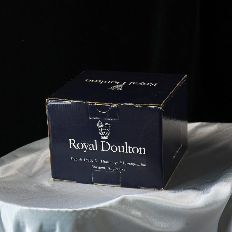 Może przedstawiać: Granatowe kartonowe pudełko z białym napisem "Royal Doulton". Pudełko zawiera r&oacute;wnież tekst "Depuis 1815, Un Hommage &agrave; l'Imagination Burslem, Angleterre" oraz logo korony. Pudełko stoi na białej satynowej powierzchni.