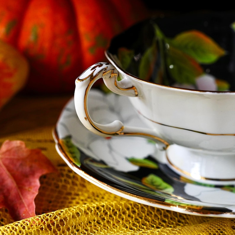 May include: A white teacup and saucer set with gold trim. The cup handle has an ornate design. The saucer features a black background with white floral and green leaf accents. The set is placed on a yellow textured surface.