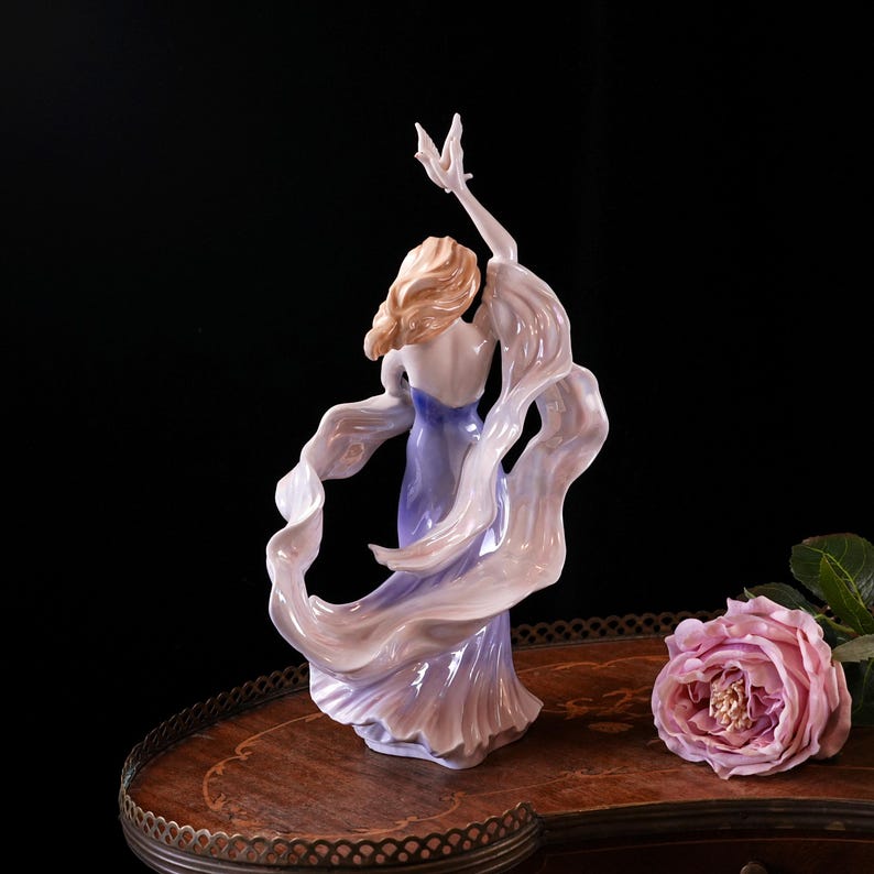 May include: A porcelain figurine of a woman in a flowing gown, with her arms raised. The dress is a gradient of lavender and white, with a touch of blue. The woman has blonde hair and is posed in a graceful dance.