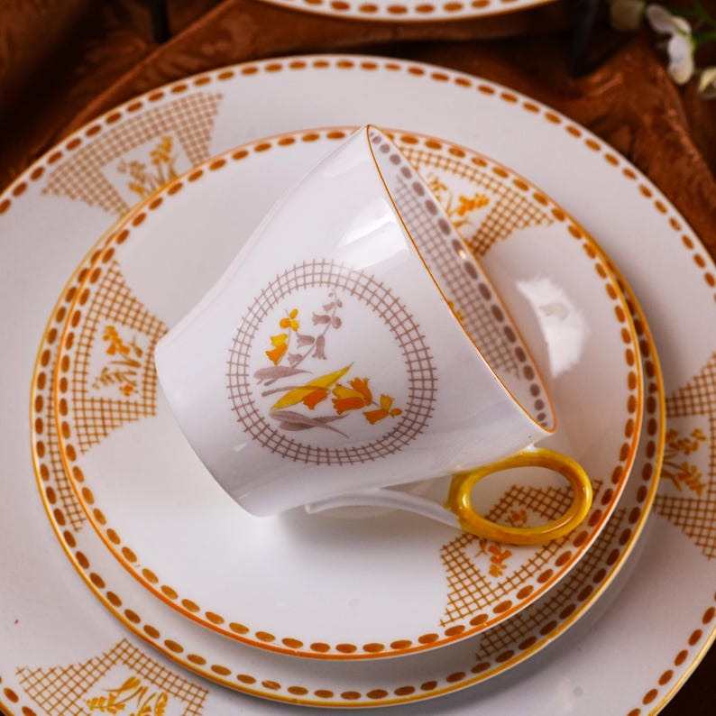 Op de afbeelding: Een witte theekop met een gele oor rust op een bijpassende schotel en bord. De set heeft een gouden rand en een herhalend patroon van oranje en bruine bloemmotieven. De theekop en schotel maken deel uit van een grotere servies set.