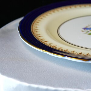 Pode incluir: Um close-up de um prato decorativo com um centro creme e uma borda azul escura. A borda tem uma borda ondulada e &eacute; adornada com ouro. O prato est&aacute; sobre uma superf&iacute;cie branca.