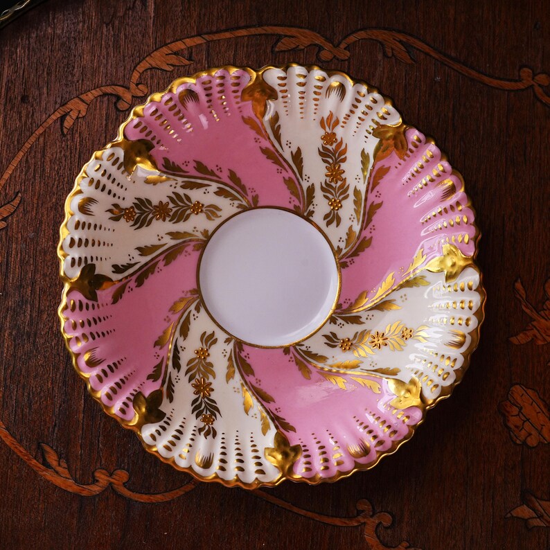 May include: A decorative, antique porcelain saucer with a scalloped edge. The saucer features a pink and white design with gold floral accents. The center is white, and the rim is edged with gold. The saucer is set on a dark wood surface.
