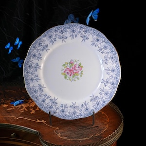 Puede incluir: Un plato redondo de cer&aacute;mica blanca con borde festoneado, que presenta un patr&oacute;n floral azul alrededor del borde y un grupo central de rosas rosadas con hojas verdes. El plato se exhibe en un soporte sobre un fondo oscuro con mariposas azules.