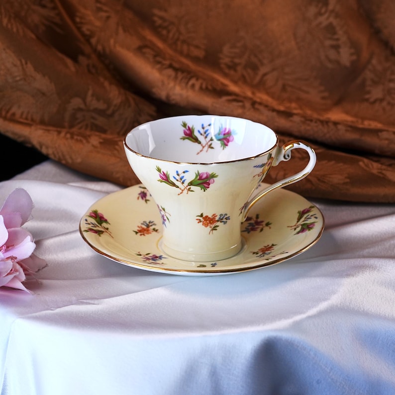 Op de afbeelding: Een vintage theekop en schotel set. De cr&egrave;me-kleurige porseleinen kop en schotel zijn versierd met bloemmotieven in roze, blauwe en oranje tinten. Gouden randen accentueren de randen van de kop en schotel, wat een vleugje elegantie toevoegt.