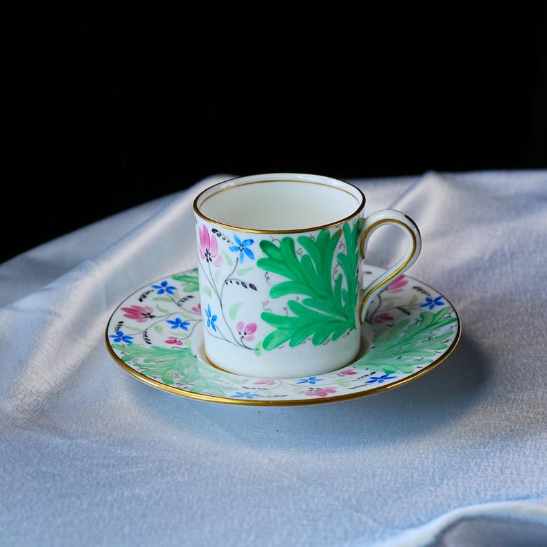 May include: A white porcelain teacup and saucer set featuring a hand-painted botanical pattern of large green acanthus leaves, small pink flowers, and tiny blue blossoms. The rim of the cup and the edge of the saucer are finished with a thin gold trim.