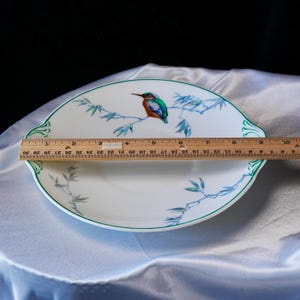 May include: White oval serving platter with a green rim and a painted bird and bamboo design. The platter measures approximately 10 inches in length. The bird is perched on a branch with blue leaves.