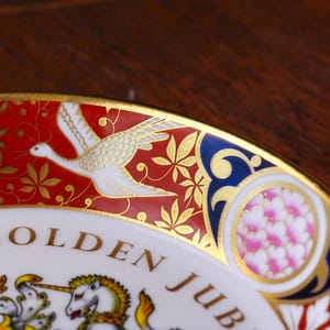 May include: A decorative porcelain plate featuring a red border with gold leaf crane motifs and a blue accent panel with pink floral patterns. The white center displays the text GOLDEN JUB and a partial illustration of a unicorn with gold details.