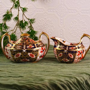 May include: A porcelain tea set with a teapot and creamer. The set features a navy blue, gold, red, and white floral design. The teapot has a lid and gold accents on the handle and spout. The creamer has a gold handle.