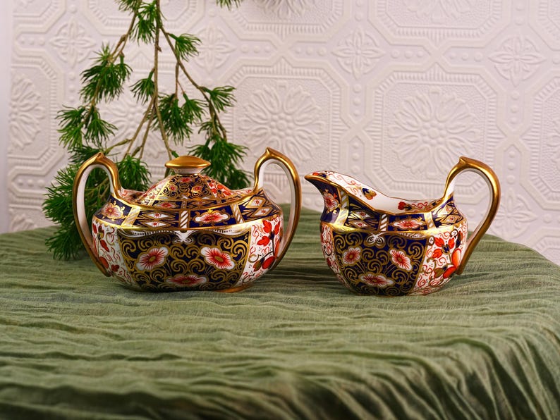 May include: A porcelain tea set with a teapot and creamer. The set features a navy blue, gold, red, and white floral design. The teapot has a lid and gold accents on the handle and spout. The creamer has a gold handle.