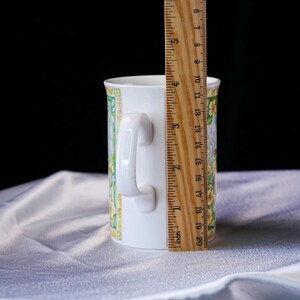 Puede incluir: Una taza de cer&aacute;mica blanca con un colorido estampado floral con detalles en amarillo y verde. Una regla de madera est&aacute; colocada verticalmente contra el lado de la taza, indicando una altura de unos 10 cm desde la base hasta el borde.